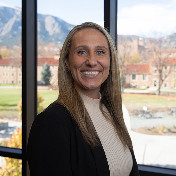 Aimee Mandolini - woman with blond hair standing in front of a window wearing a beige turtleneck sweater and black blazer.