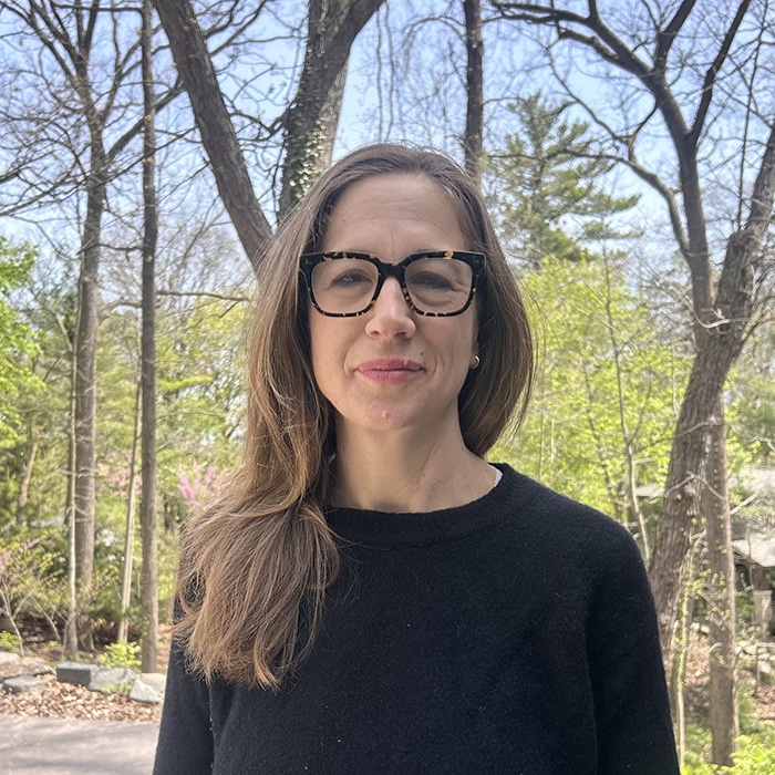 Caitlin Shamberg - A woman with light brown hair and glasses standing outdoors in a wooded area wearing a black sweater