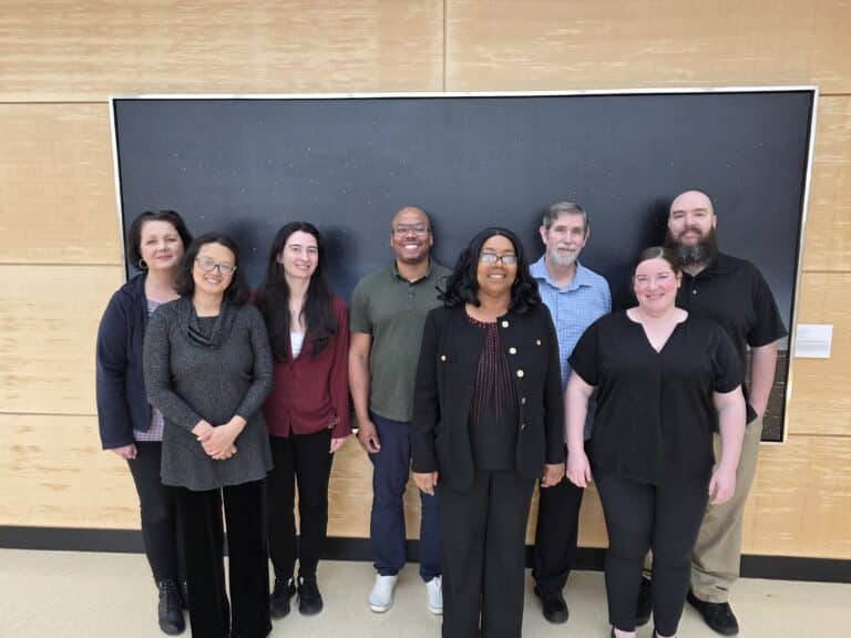 Eight staff members from the PhD and Research Support office are smiling in front of a black board
